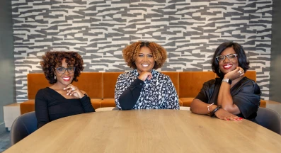 Three people sit at a wooden conference table in a modern office. Behind them is a patterned gray‑and‑white wall and a long orange‑brown cushioned bench. All three are smiling, wearing glasses, and dressed in stylish business‑casual clothing. Bright, even lighting creates a polished, collaborative atmosphere.